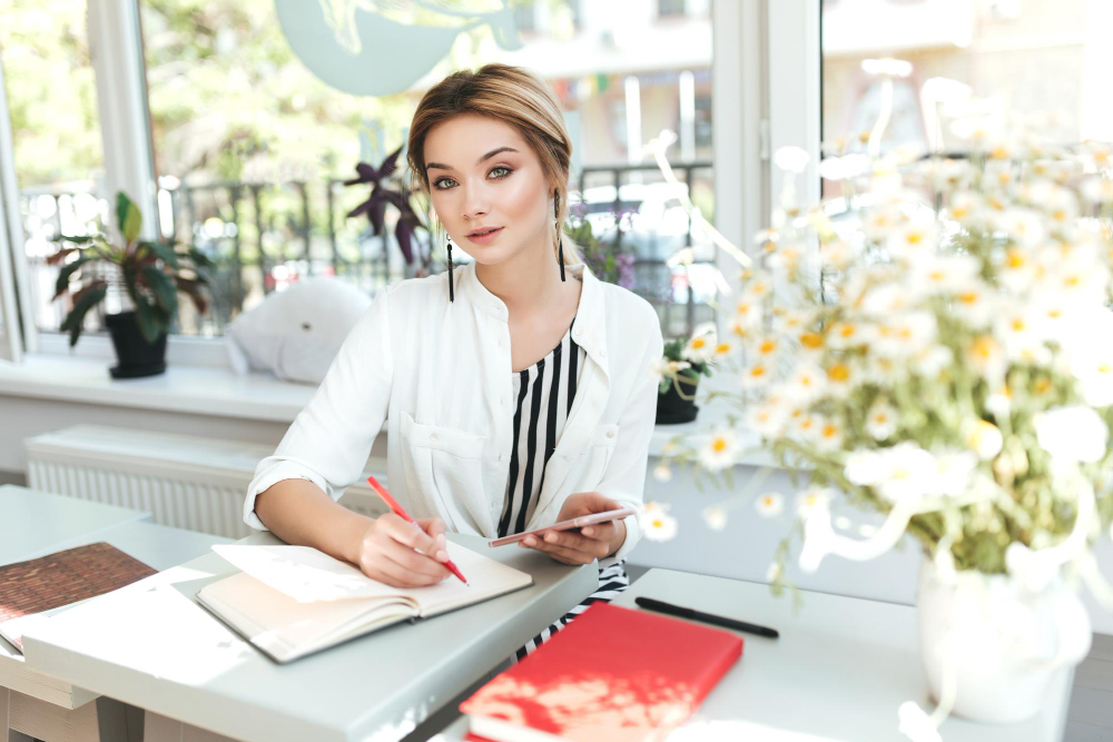 portrait-beautiful-girl-sitting-restaurant-with-notebook-looking-camera-pretty-girl-with-blond-hair-holding-mobile-phone-hand-writing-notes-her-notebook-cafe